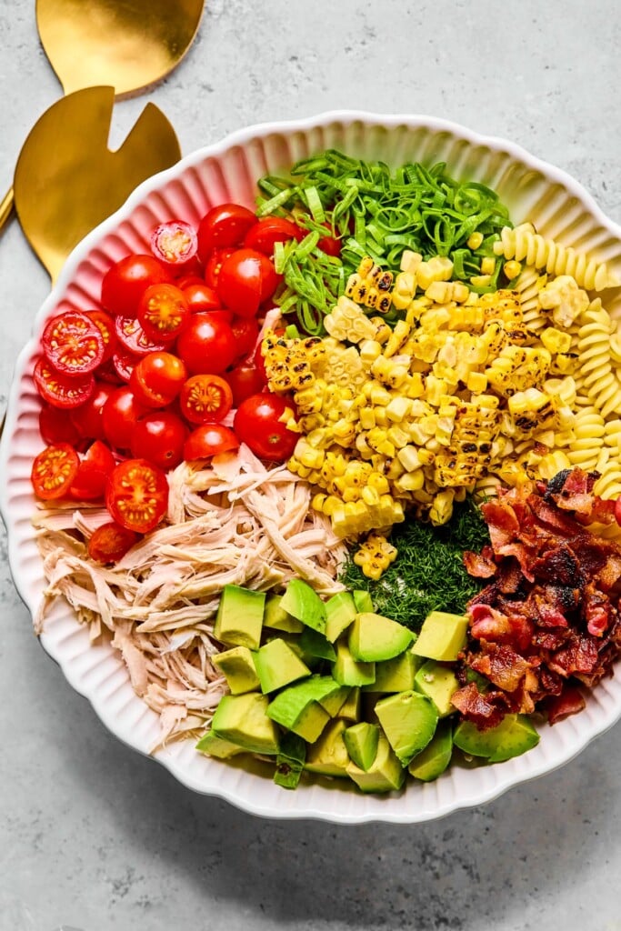 Large bowl of cooked pasta, charred corn that has been cut off the cob, pieces of bacon, fresh dill, diced avocado, shredded chicken, halved cherry tomatoes, and sliced green onions before it is mixed together. Next to the bowl are gold mixing spoons.