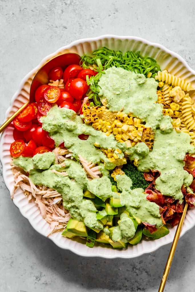 Large bowl of cooked pasta, charred corn that has been cut off the cob, pieces of bacon, fresh dill, diced avocado, shredded chicken, halved cherry tomatoes, and sliced green onions with jalapeño ranch poured on top of it.