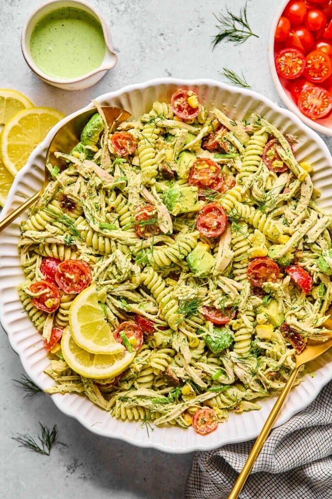 Chicken bacon pasta salad tossed with jalapeño ranch in a large white bowl. The pasta salad is garnished with fresh dill and lemon slices and there are two gold spoons in the bowl. Around the bowl are lemon slices, a cup of jalapeño ranch, a bowl of halved cherry tomatoes, and a kitchen towel.