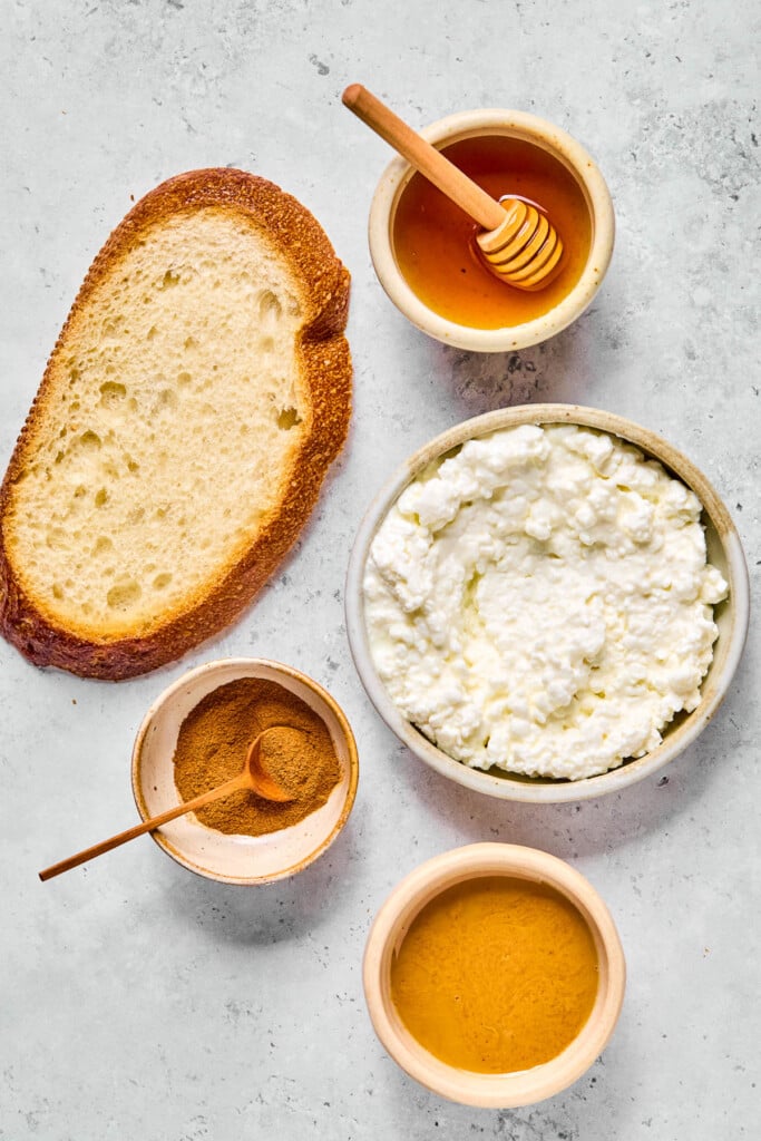 Grey concrete counter with a small bowl of honey with a honey dipper in it, a bowl of cottage cheese, a small bowl of peanut butter, a small bowl of cinnamon with a small spoon in it, and a piece of bread on it.