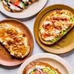 Four plates of cottage cheese toast with different toppings on each one. One has sliced tomato, balsamic glaze, and basil on it. Another has smashed avocado, chili oil, and honey on it. The third one has sliced cucumber, smoked salmon, red onion, dill, and everything but the bagel seasoning on it. The last one has peanut butter, honey, and cinnamon on it.