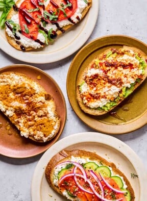 Four plates of cottage cheese toast with different toppings on each one. One has sliced tomato, balsamic glaze, and basil on it. Another has smashed avocado, chili oil, and honey on it. The third one has sliced cucumber, smoked salmon, red onion, dill, and everything but the bagel seasoning on it. The last one has peanut butter, honey, and cinnamon on it.