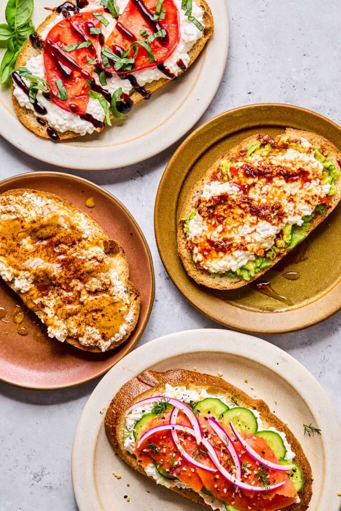Four plates of cottage cheese toast with different toppings on each one. One has sliced tomato, balsamic glaze, and basil on it. Another has smashed avocado, chili oil, and honey on it. The third one has sliced cucumber, smoked salmon, red onion, dill, and everything but the bagel seasoning on it. The last one has peanut butter, honey, and cinnamon on it.