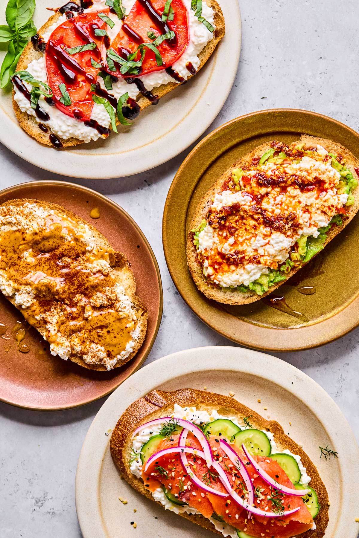 Four plates of cottage cheese toast with different toppings on each one. One has sliced tomato, balsamic glaze, and basil on it. Another has smashed avocado, chili oil, and honey on it. The third one has sliced cucumber, smoked salmon, red onion, dill, and everything but the bagel seasoning on it. The last one has peanut butter, honey, and cinnamon on it.