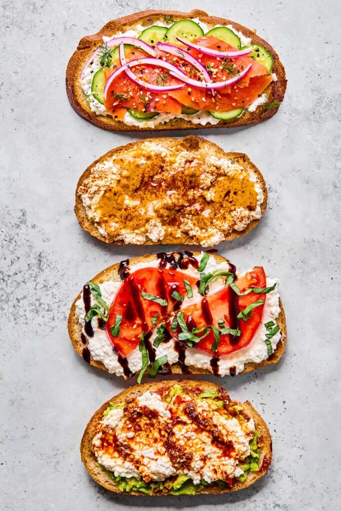 Four pieces of cottage cheese on toast. One has smoked salmon and veggies on it. Another has peanut butter and honey on it. The third one has sliced tomato and balsamic on it. And the fourth one has avocado and chili oil on it.