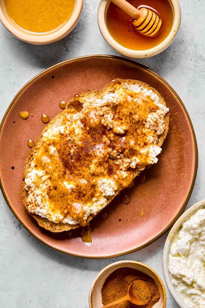 A plate with a piece of toast topped with cottage cheese, peanut butter, honey, and cinnamon on it. Around the plate is a bowl of peanut butter, a small bowl of honey with a honey dipper in it, a bowl of cottage cheese, and a small bowl of cinnamon.