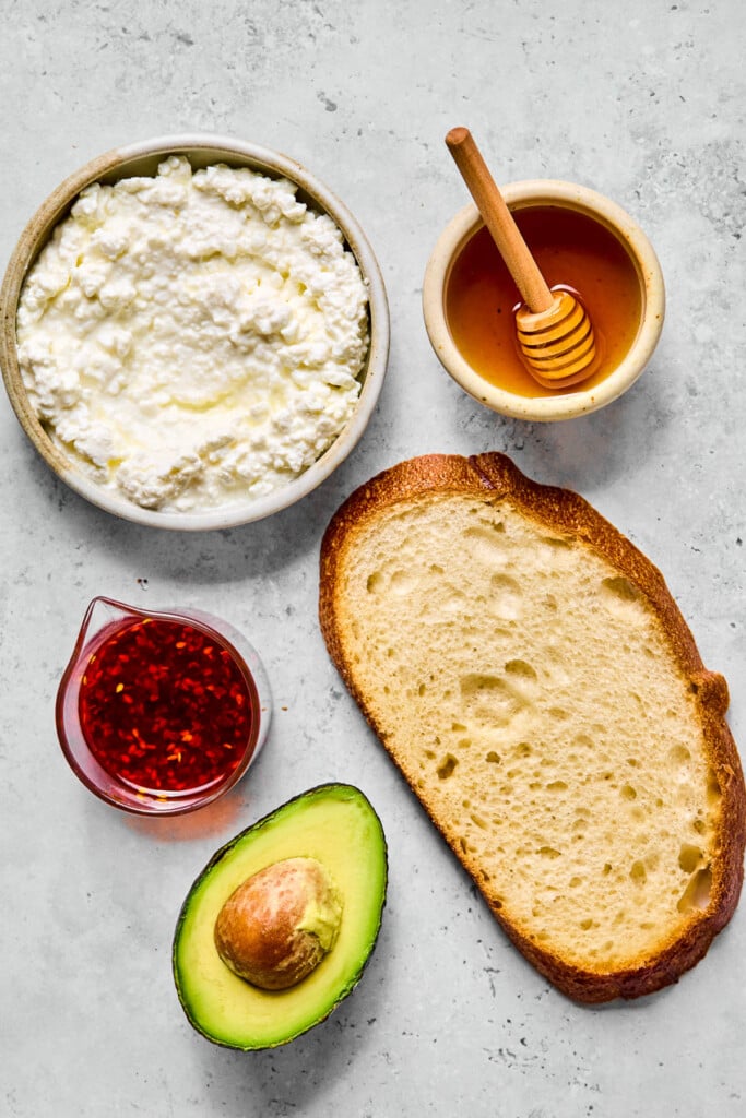 Grey concrete counter with a small bowl of honey with a honey dipper in it, a piece of bread, half an avocado, a small cup of chili oil, and a bowl of cottage cheese on it.
