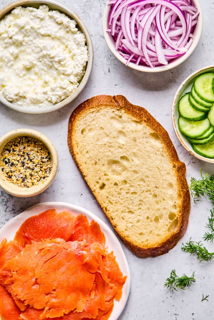 Grey concrete counter with a bowl of sliced red onions, a bowl of sliced cucumber, a piece of bread, springs of fresh dill, a plate of smoked salmon, a small bowl of everything but the bagel seasoning, and a bowl of cottage cheese on it.