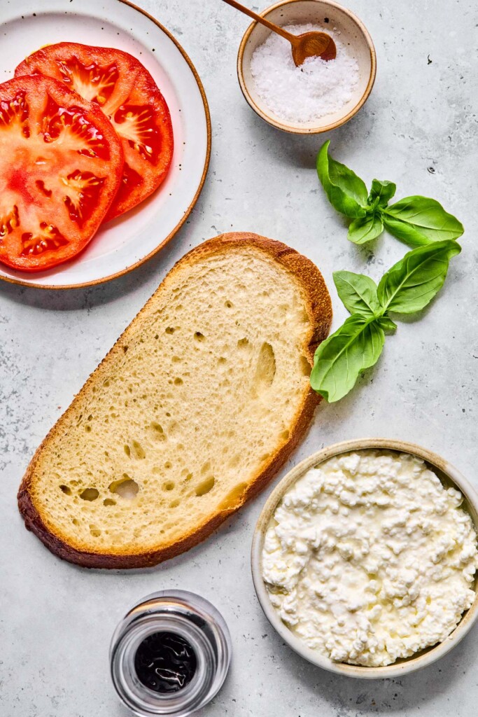 Grey concrete counter with a small bowl of flaky salt with a small spoon in it, fresh basil leaves, a bowl of cottage cheese, a small dish with balsamic glaze, a piece of bread, and a plate with some tomato slices on it.