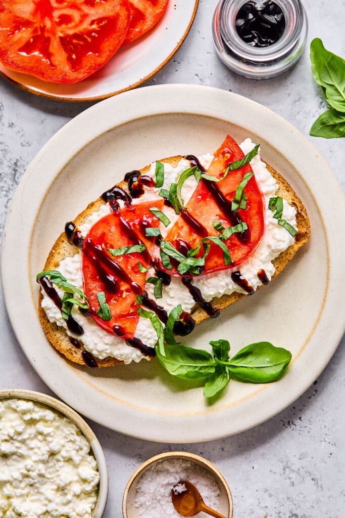 Cottage cheese tomato toast topped with balsamic glaze and basil on a white plate. Around the plate is a small jar with balsamic glaze, fresh basil, a small bowl of salt, a bowl of cottage cheese, and a plate with tomatoes on it.