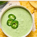 A bowl of jalapeno ranch garnished with sliced jalapenos. The bowl is on a plate that is filled with tortilla chips.