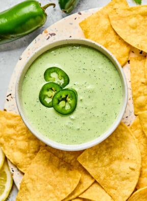 A bowl of jalapeno ranch garnished with sliced jalapenos. The bowl is on a plate that is filled with tortilla chips. Next to the plate are some whole jalapenos.