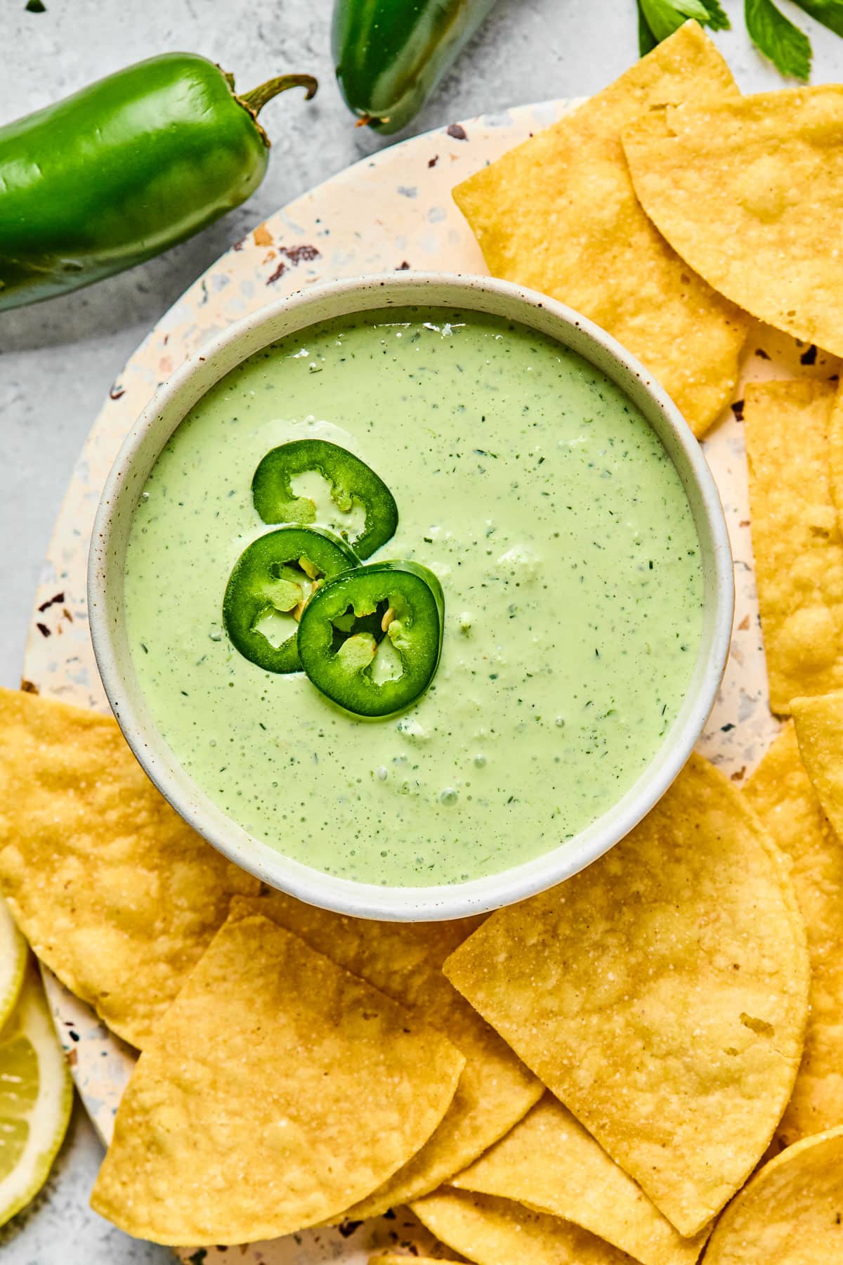 A bowl of jalapeno ranch garnished with sliced jalapenos. The bowl is on a plate that is filled with tortilla chips. Next to the plate are some whole jalapenos.