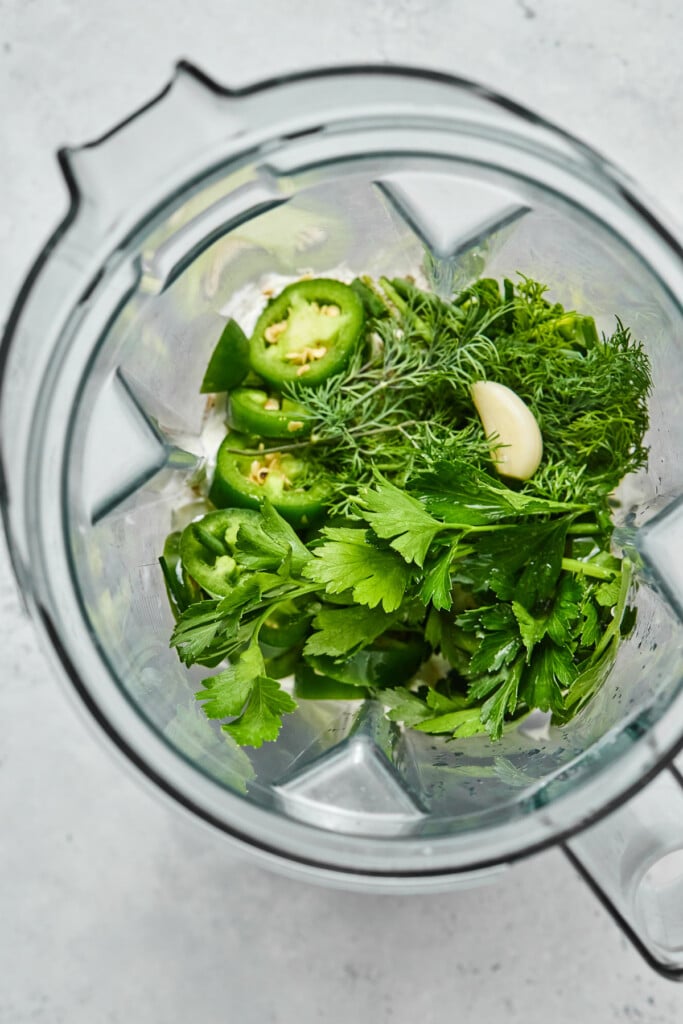 Overhead shot of a blender filled with greek yogurt, a bunch of fresh herbs, sliced jalapenos, and a garlic clove.