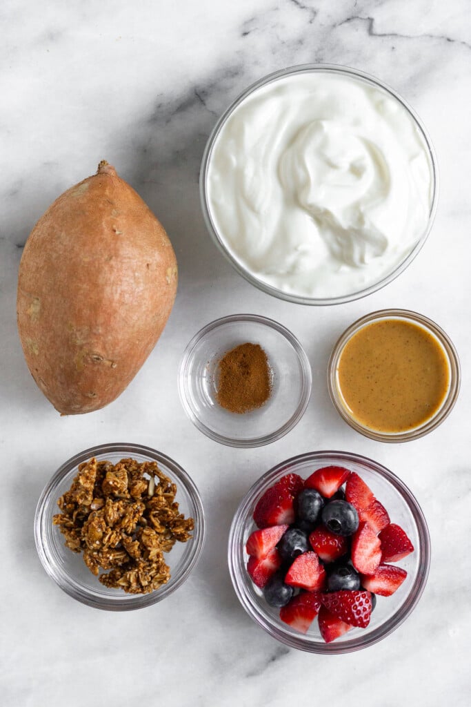 White marble counter with a bowl of greek yogurt, a small bowl of peanut butter, a small bowl of cinnamon, a bowl of blueberries and diced strawberries, a bowl of granola, and a small uncooked sweet potato on it.