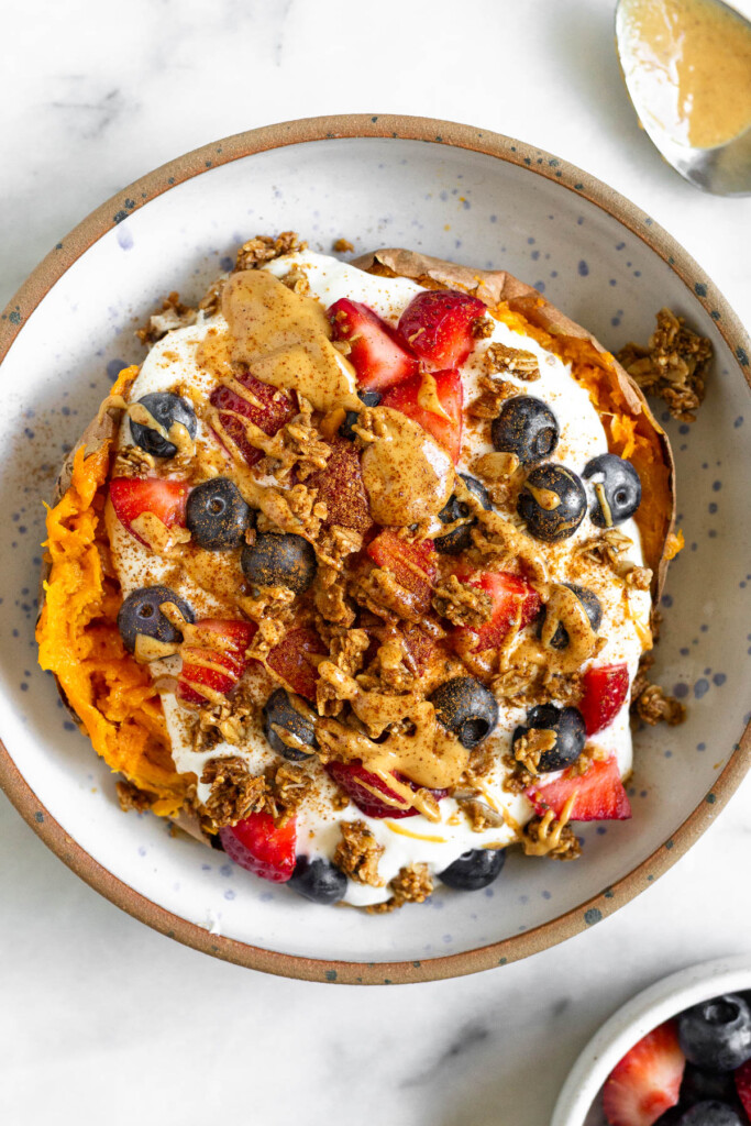 Greek yogurt stuffed a cooked sweet potato topped with berries, granola, nut butter, and cinnamon in a bowl. Next to the bowl is a bowl of berries and a spoon with peanut butter on it.
