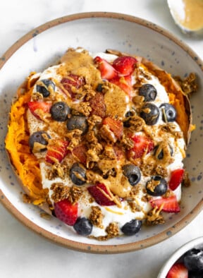 Sweet potato breakfast bowl with greek yogurt, berries, nut butter, granola, and topped with cinnamon. Next to the bowl is a bowl of berries and a spoon with peanut butter on it.