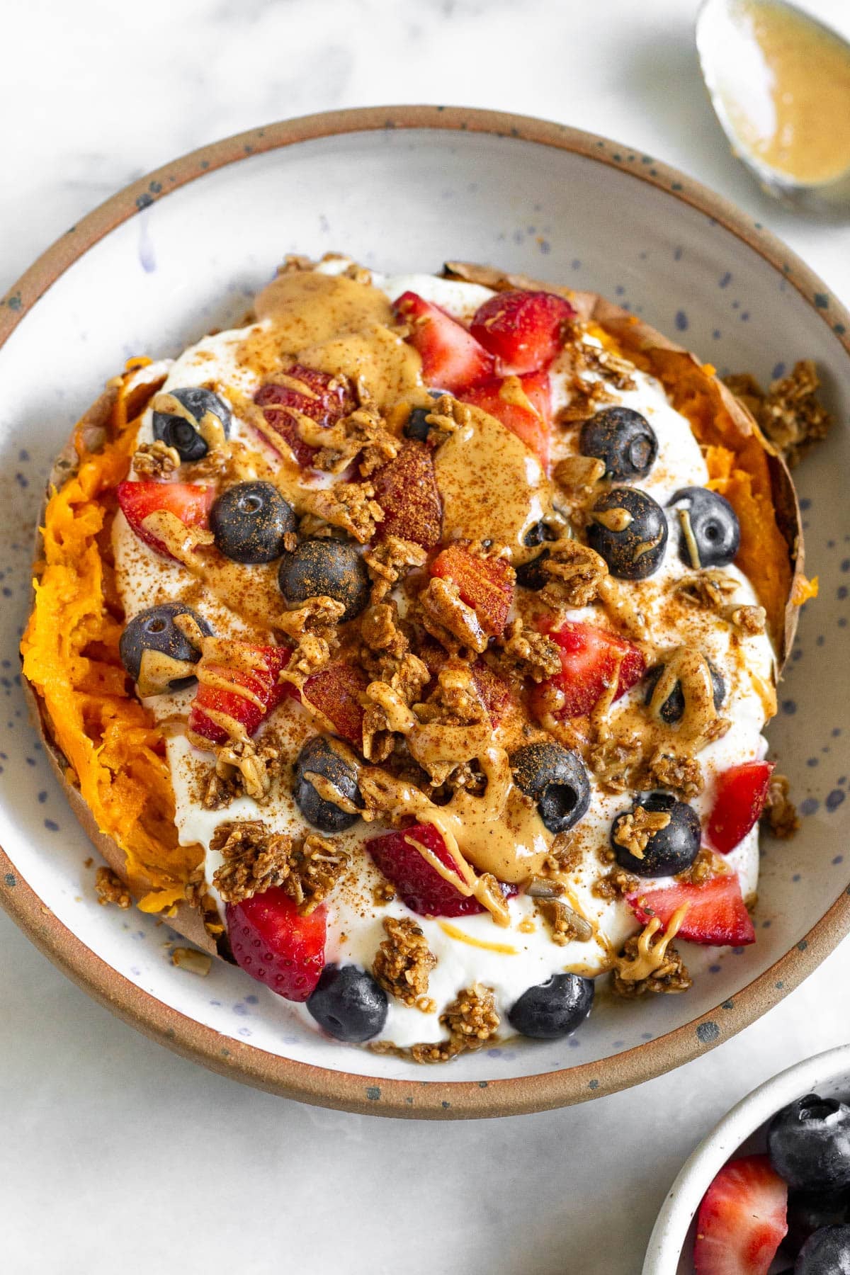 Sweet potato breakfast bowl with greek yogurt, berries, nut butter, granola, and topped with cinnamon. Next to the bowl is a bowl of berries and a spoon with peanut butter on it.
