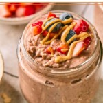 Jar of chocolate overnight oats topped with diced strawberries, blueberries, and a drizzle of peanut butter. Around the jar are some rolled oats, a blueberry, a bowl of diced strawberries, and another jar of overnight oats.