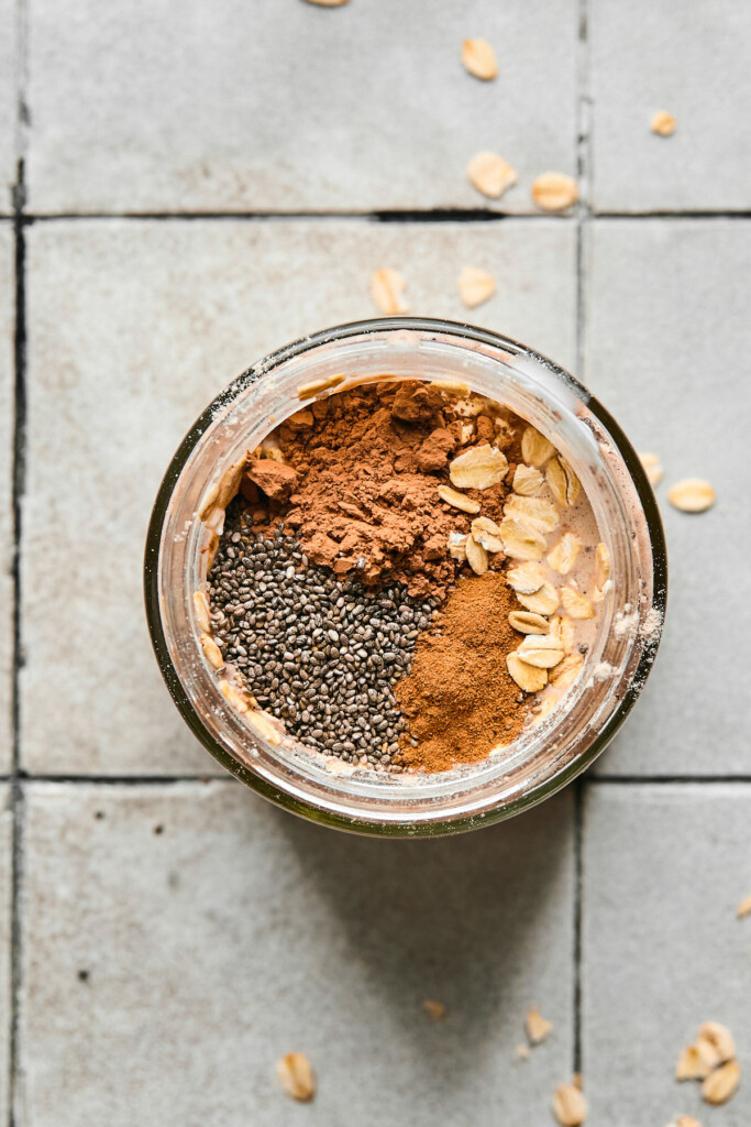 An overhead shot of a jar with milk, rolled oats, cinnamon, chia seeds, and cocoa powder before it is mixed together.