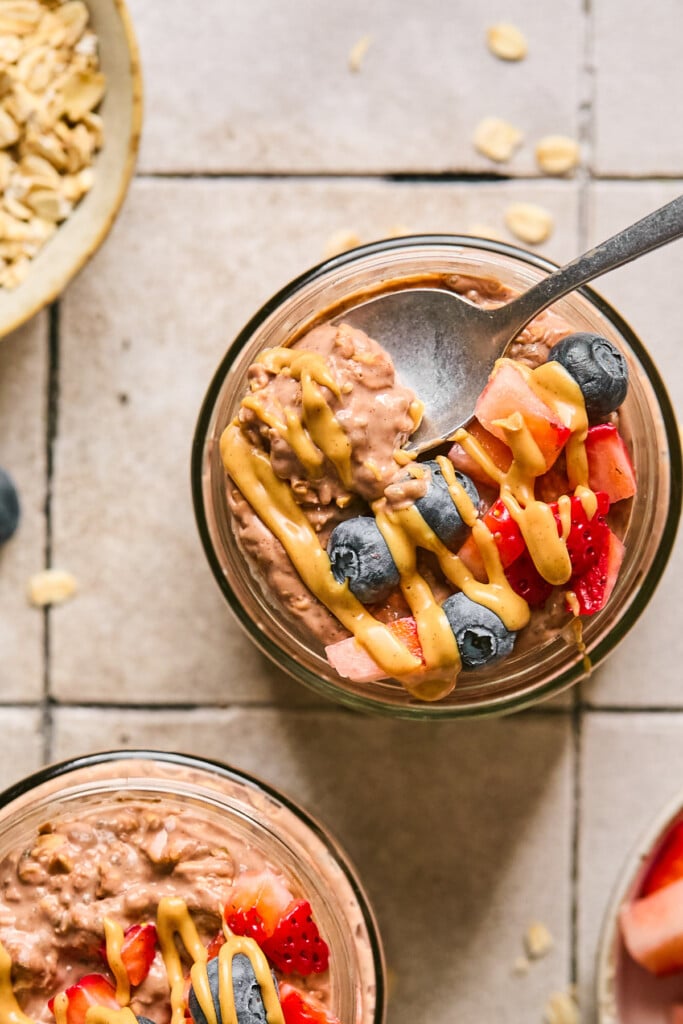 Overhead shot of a jar of chocolate overnight oats topped with blueberries, diced strawberries, and a drizzle of peanut butter. A spoon is overtop the jar with a scoop of overnight oats on it. Around the jar is a bowl of diced strawberries, another jar of overnight oats, and a bowl of rolled oats.