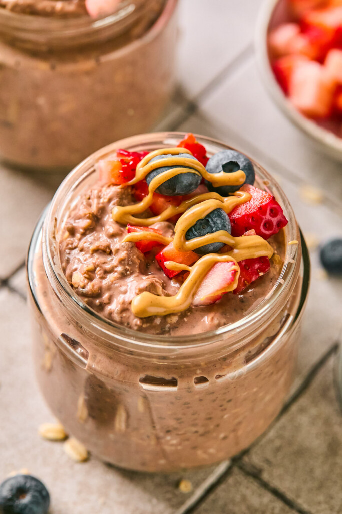 A jar of chocolate protein overnight oats topped with diced strawberries, blueberries, and a drizzle of peanut butter. Around the jar are some blueberries, a bowl of diced strawberries, and another jar of overnight oats.