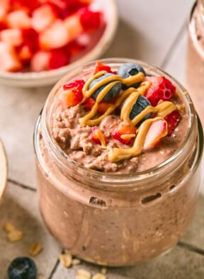 Jar of chocolate overnight oats topped with diced strawberries, blueberries, and a drizzle of peanut butter. Around the jar are some rolled oats, a blueberry, a bowl of diced strawberries, and another jar of overnight oats.