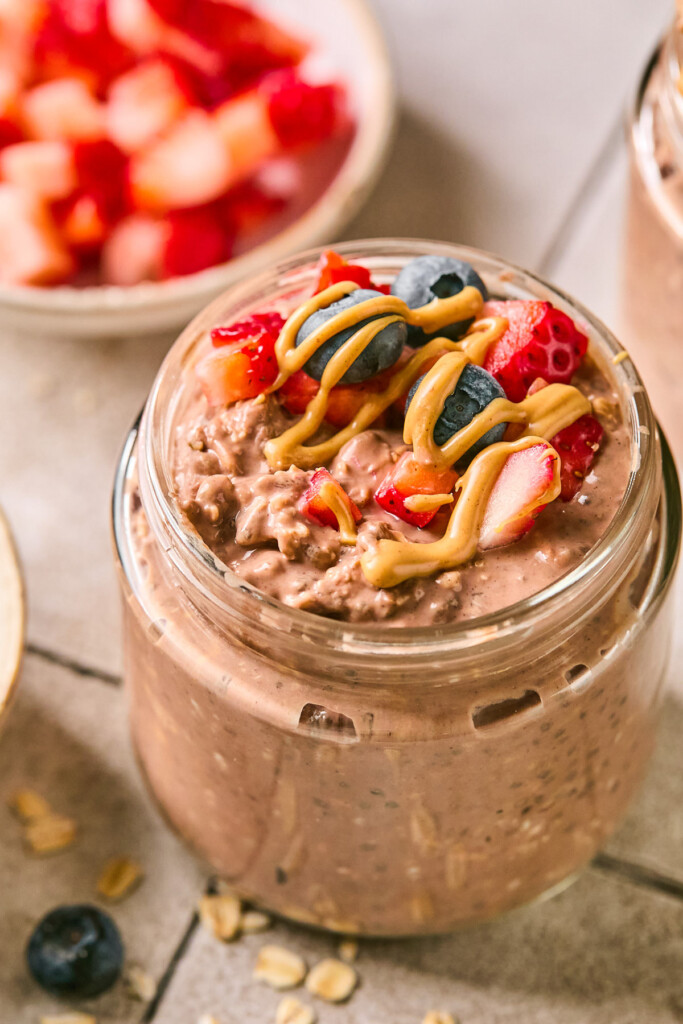 Jar of chocolate overnight oats topped with diced strawberries, blueberries, and a drizzle of peanut butter. Around the jar are some rolled oats, a blueberry, a bowl of diced strawberries, and another jar of overnight oats.