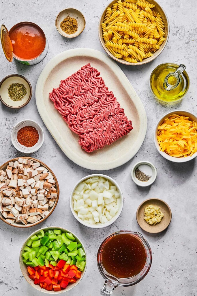 Grey concrete counter with a bowl of uncooked pasta, a small pitcher of olive oil, a bowl of shredded cheese, a small bowl of salt and pepper, a small bowl of minced garlic, a measuring cup of bone broth, a bowl of diced onion, a bowl of diced red and green bell peppers, a bowl of diced mushrooms, a plate of pound of raw ground beef, a small bowl of chili powder, a small bowl of dried oregano, a can of red enchilada sauce, and a small bowl of cumin on it.
