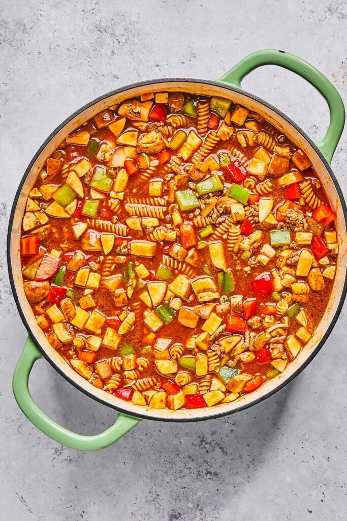 Large enameled pan filled with a mixture of ground meat, diced veggies, uncooked pasta, and red sauce before it is cooked.