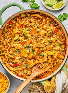 Overhead shot of enchilada pasta in a large pot topped with melted cheese and cilantro. A wooden spoon is also in the pot. Around the pot is a a plate of lime wedges, some sprigs of fresh cilantro, a tan kitchen towel, and a bowl of shredded cheddar cheese.