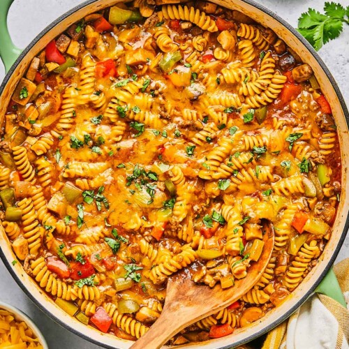 Overhead shot of enchilada pasta in a large pot topped with melted cheese and cilantro. A wooden spoon is also in the pot. Around the pot is a a plate of lime wedges, some sprigs of fresh cilantro, a tan kitchen towel, and a bowl of shredded cheddar cheese.