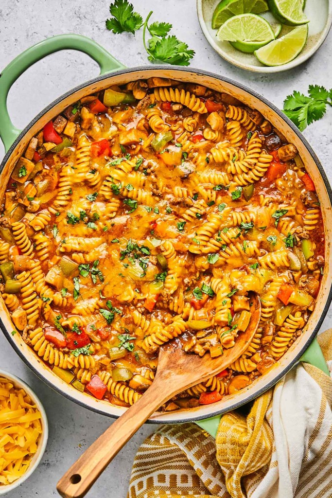 Overhead shot of enchilada pasta in a large pot topped with melted cheese and cilantro. A wooden spoon is also in the pot. Around the pot is a a plate of lime wedges, some sprigs of fresh cilantro, a tan kitchen towel, and a bowl of shredded cheddar cheese.