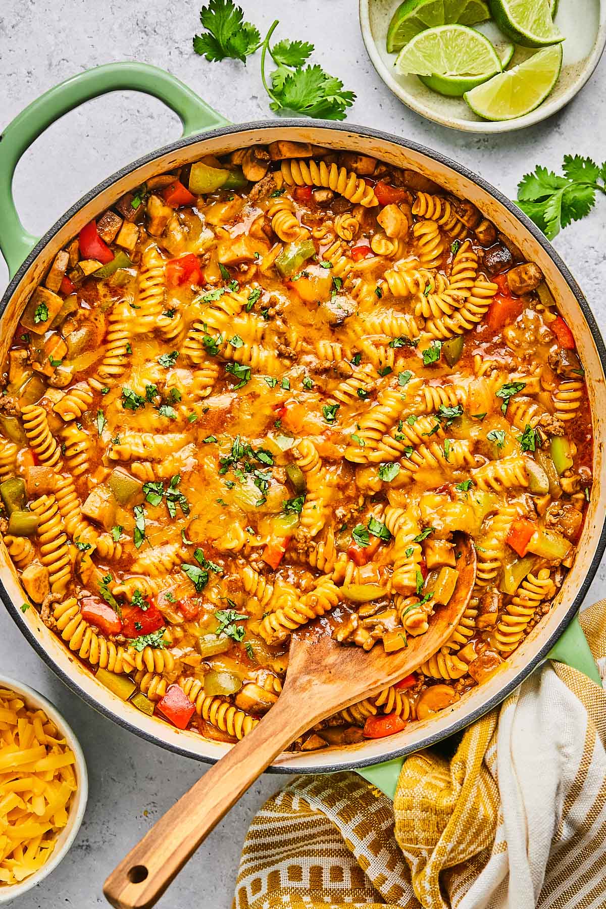Overhead shot of enchilada pasta in a large pot topped with melted cheese and cilantro. A wooden spoon is also in the pot. Around the pot is a a plate of lime wedges, some sprigs of fresh cilantro, a tan kitchen towel, and a bowl of shredded cheddar cheese.