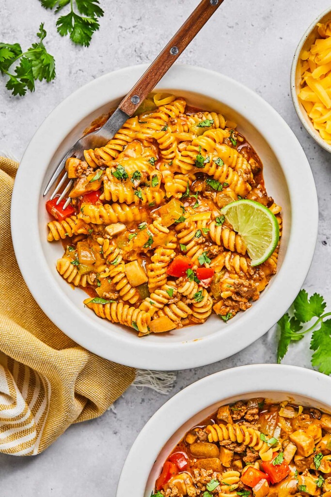 Two bowls of enchilada pasta topped with cilantro. One of the bowls is garnished with a lime wedge and has a fork in it. Around the bowls is fresh cilantro, a tan kitchen towel, and bowl of shredded cheese.