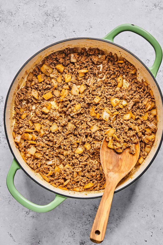 Large enameled pan filled sautéed onions and seasoning ground beef. There is a wooden spoon in the pan as well.