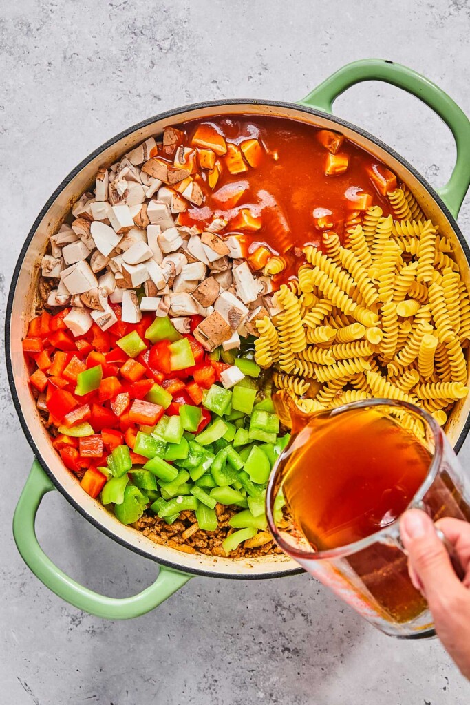 Overhead shot of a large enameled pan filled with uncooked pasta, seasoned ground beef, diced green and red bell peppers, and diced mushrooms, with red enchilada sauce being poured overtop.