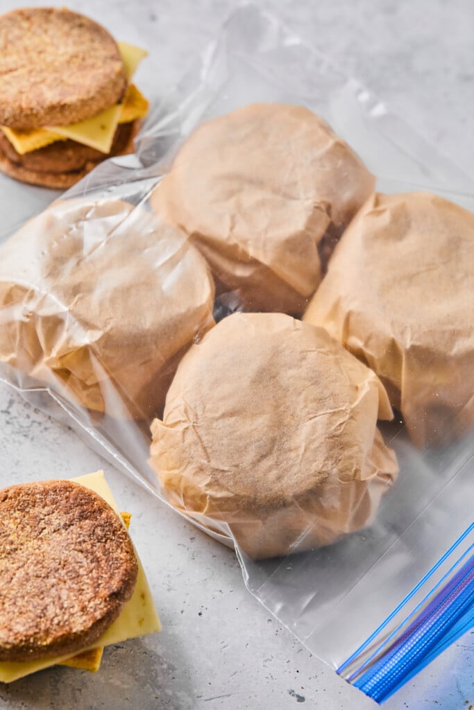 Frozen breakfast sandwiches wrapped in parchment paper in a large plastic bag. Next to the bag are two sandwiches that haven't been wrapped up yet.