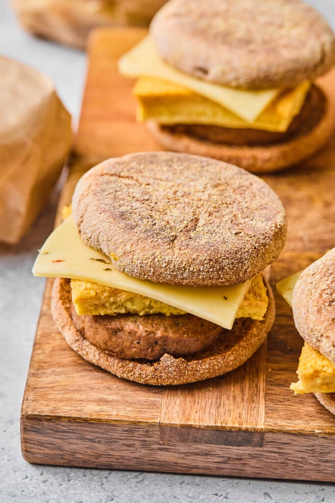 A few healthy breakfast sandwiches with eggs, chicken sausage, and cheese in an English muffin on a wooden cutting board.