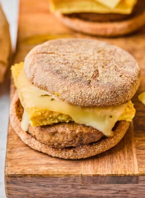 Freezer breakfast sandwich with sausage, egg, and cheese on an English muffin on a wooden cutting board. There are some sandwiches in the background.