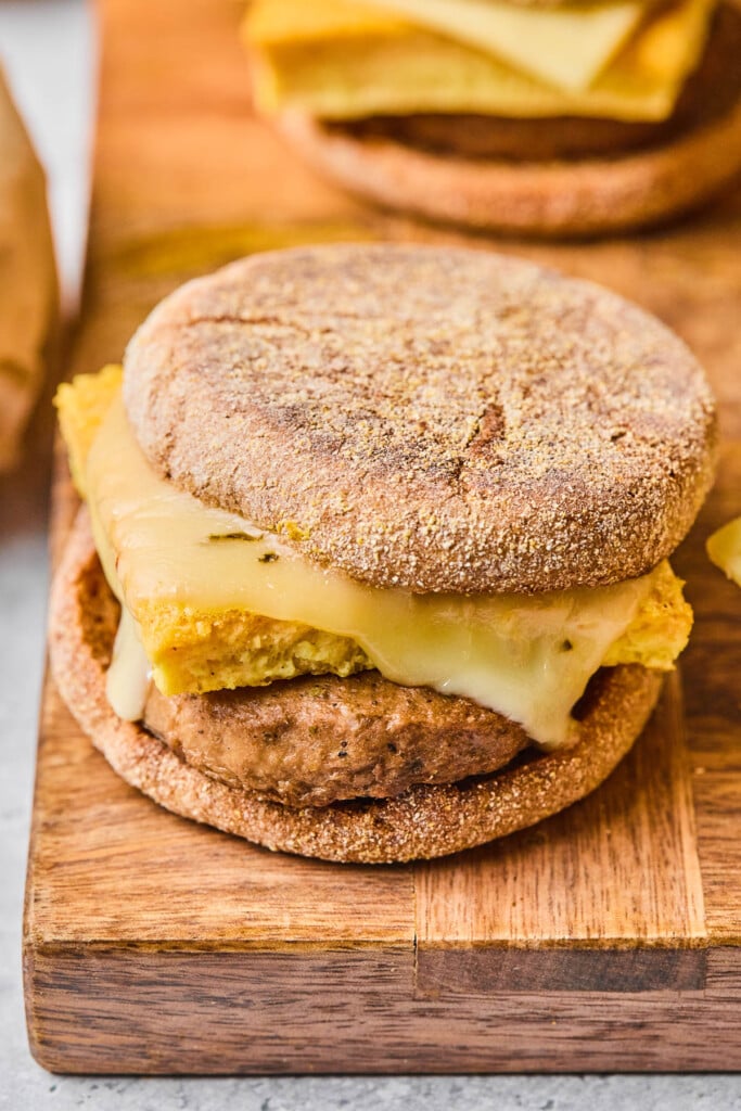 Freezer breakfast sandwich with sausage, egg, and cheese on an English muffin on a wooden cutting board. There are some sandwiches in the background.