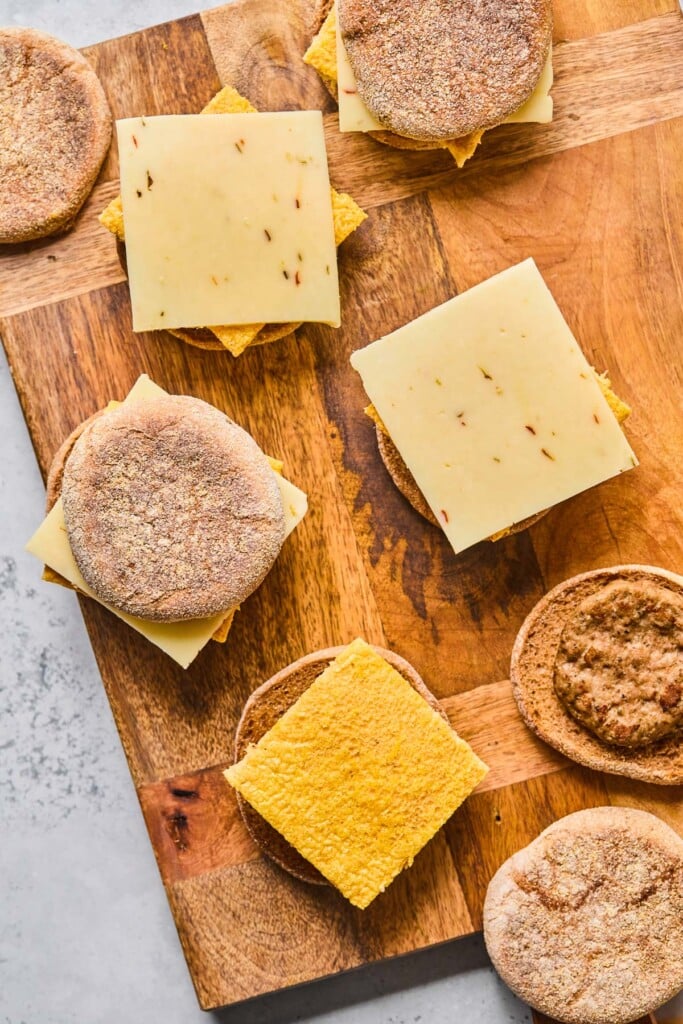 Overhead shot of breakfast sandwiches being assembled on a wooden cutting board. Some have the bottom of an English muffin with a chicken sausage, some have the bottom of an English muffin with a piece of baked egg, some have the bottom of an English muffin with a piece of baked egg and a slice of cheese, and some are fully assembled with the top of the English muffin on the sandwich.