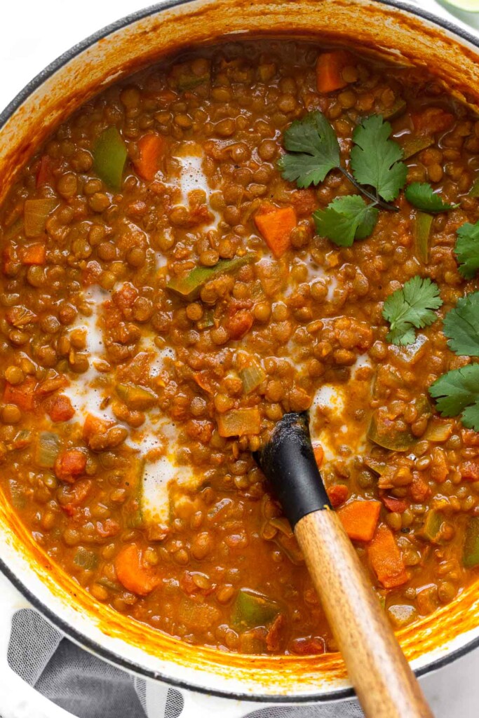 A large white dutch oven with coconut curry lentil soup garnished with cilantro and a drizzle of coconut milk. A spoon is also in the pot.