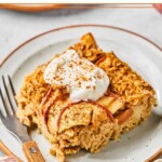 A piece of apple cinnamon baked oatmeal on a plate. It is garnished with greek yogurt and cinnamon and a fork is also on the plate. Around the plate is a bowl of walnuts and a red and white checkered kitchen towel.