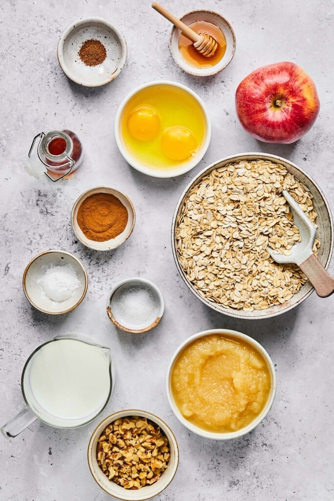 A small bowl of honey with a honey dipper in it, a red apple, a bowl of rolled oats with a scooper in it, a bowl of unsweetened applesauce, a bowl of chopped walnuts, a measuring cup of milk, a small bowl of salt, a small bowl of baking powder, a small bowl of cinnamon, a small jar of vanilla extract, a bowl with two cracked eggs in it, and a small bowl of nutmeg on a grey concrete countertop.