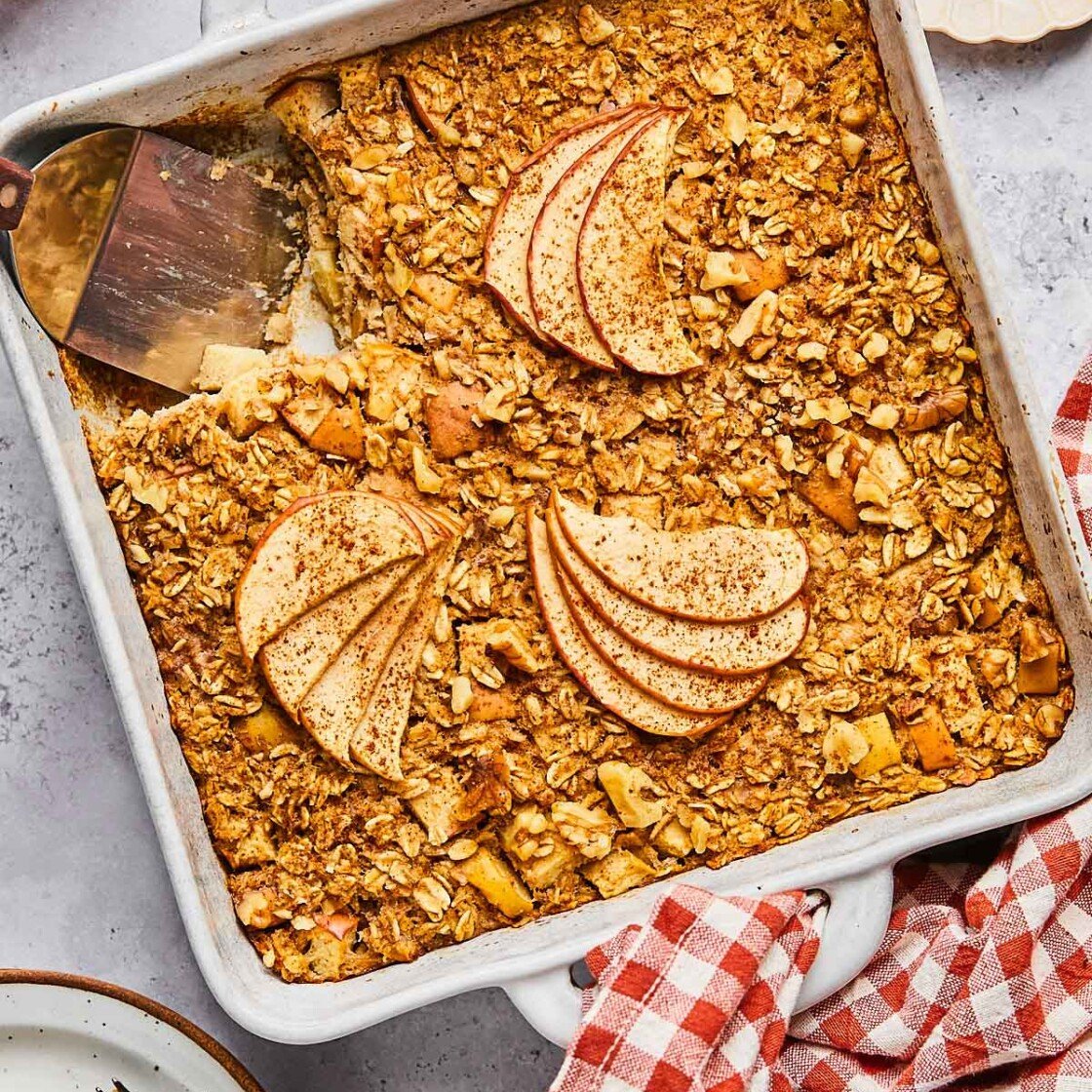 A plan of apple cinnamon baked oatmeal with a piece cut out of it. A spatula is in the pan where the pieces is gone. Around the pan is a a plate of chopped walnuts, a red and white checkered towel, a plate with a fork and a piece of the baked oatmeal on it, and some red apples.