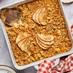 A plan of apple cinnamon baked oatmeal with a piece cut out of it. A spatula is in the pan where the pieces is gone. Around the pan is a a plate of chopped walnuts, a red and white checkered towel, a plate with a fork and a piece of the baked oatmeal on it, and some red apples.