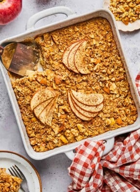 A plan of apple cinnamon baked oatmeal with a piece cut out of it. A spatula is in the pan where the pieces is gone. Around the pan is a a plate of chopped walnuts, a red and white checkered towel, a plate with a fork and a piece of the baked oatmeal on it, and some red apples.