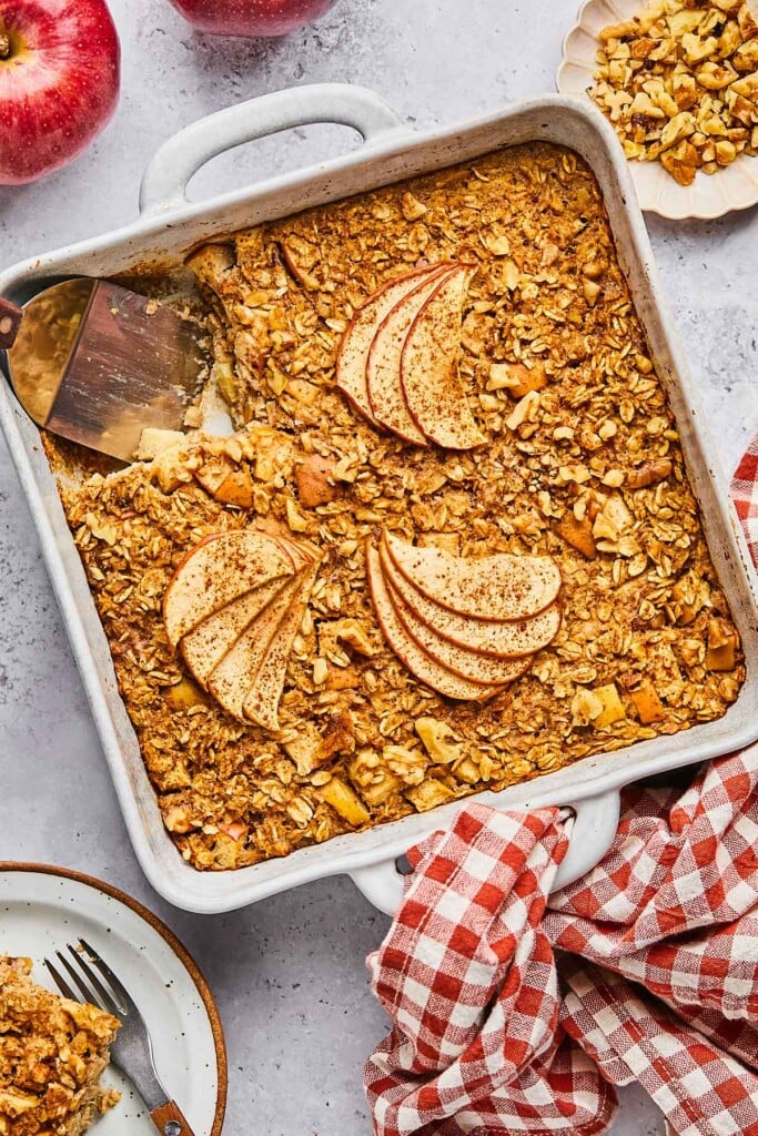 A plan of apple cinnamon baked oatmeal with a piece cut out of it. A spatula is in the pan where the pieces is gone. Around the pan is a a plate of chopped walnuts, a red and white checkered towel, a plate with a fork and a piece of the baked oatmeal on it, and some red apples.