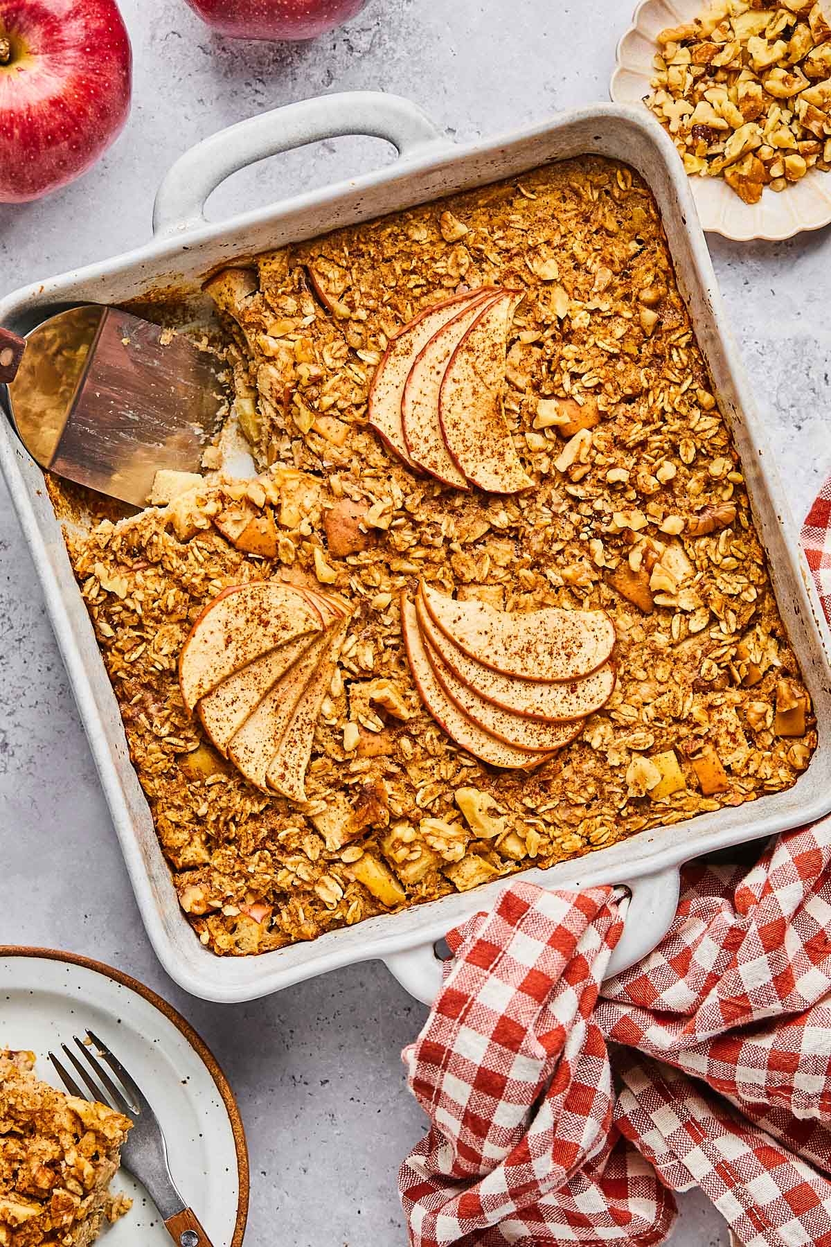 A plan of apple cinnamon baked oatmeal with a piece cut out of it. A spatula is in the pan where the pieces is gone. Around the pan is a a plate of chopped walnuts, a red and white checkered towel, a plate with a fork and a piece of the baked oatmeal on it, and some red apples.